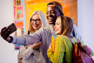 Cheerful multiethnic students taking photo with mobile phone in college. Multiracial friendship, study abroad, concept.