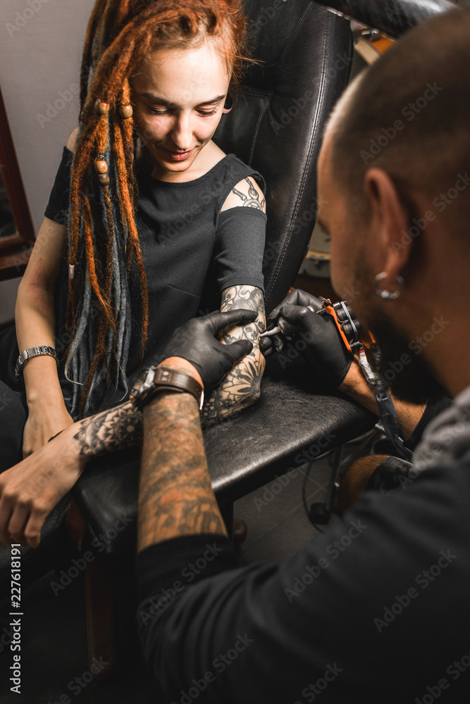 Wall mural girl with dreadlocks in a tattoo parlor. The master creates a picture on the body of a young beautiful girl. Close-up of hands and tattoo machine