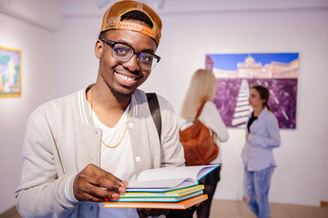 Afro-american hipster student man in eyeglasses holding colorful stack of books, smile, looking at camera. Guy in art gallery with two girls on background. Multi ethnic, study abroad, culture concept.