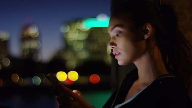 Young Woman Uses Her Smartphone At Night, Her Face Lit By Screen, City In Background