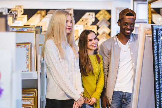 Three multi ethnic people students reflecting at mirror with expensive frame indoor at frames store, smiling. Young friendly black skinned guy and two european girls choosing a gift.