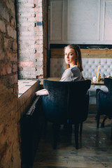 Beautiful young woman sits near big window at cafe, and looking at the camera.