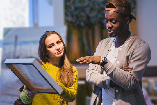 Young Hipster Couple With Different Skins Choosing A Picture In Art Gallery Or Store. Multi Ethnic Woman And Man Buying Gifts For New Housing. Friendship, Relations And Family Concept.