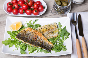 Grilled seabass fillet with arugula, lemon, tomatoes and capers. Close-up.