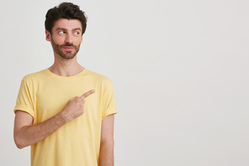 Close up of attractive young man with restrained smile, dark brown hair and beard wears yellow casual tshirt, points to the right side with finger isolated over white background