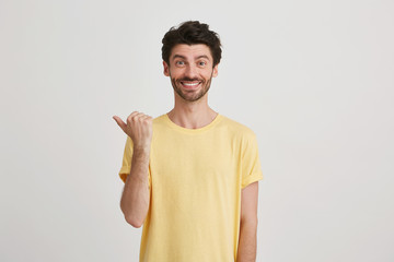 Fototapeta premium Smiling happy attractive young man with dark brown hair and beard wears yellow tshirt, points left with one thumb, isolated over white background