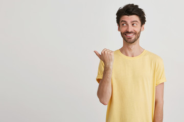 Indoor photo of smiling happy attractive young man with dark brown hair and beard wears yellow casual tshirt, points left with one thumband looks left side, isolated over white background