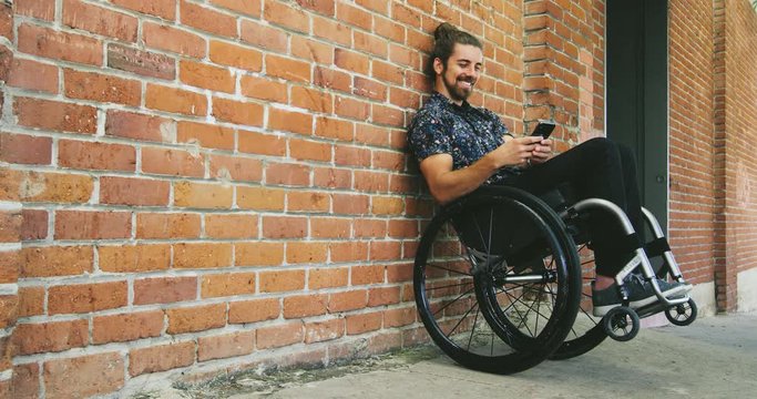 Young happy disabled man texting in wheelchair leaning against brick wall in city