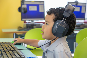 A small boy wearing oversized headphones navigates a wireless mouse in front of the computer. He...