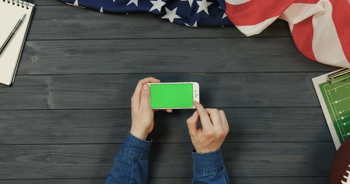View from above on the white smartphone device lying horizontally on the dark grey wooden desk with rugby ball and American flag while male hands scrolling and taping on the green screen of it. Chroma