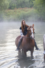 Young woman on a brown horse stays in the lake. Fog