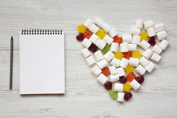 Blank notepad with pencil and heart made of candies over white wooden background, top view. From...