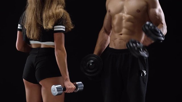 Athletic man and woman flexes their hands with dumbbells, training their biceps on a black background in studio