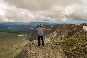 Obraz premium Guy in a gray sweatshirt with a cap is standing with his hands apart on the edge of the cliff in front of him is a colorful view of mountains lakes with glaciers. Traveler complete the route