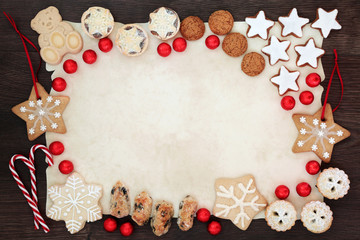 Christmas biscuit background border with delicious home made biscuits, candy canes, cakes and foil wrapped chocolates on parchment paper on rustic oak wood. Top view.