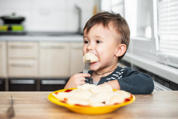 Hungry child eating dumplings in the kitchen, sitting at the table in a gray jacket