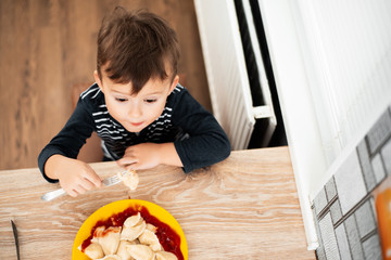 Hungry child eating dumplings in the kitchen, sitting at the table in a gray jacket