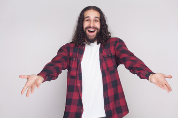Welcome, nice to see you. Surprised man with beard and black long curly hair in casual checkered shirt looking at camera with raised arms and happiness. indoor studio shot, isolated on grey background