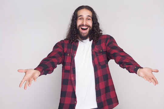 Welcome, nice to see you. Surprised man with beard and black long curly hair in casual checkered shirt looking at camera with raised arms and happiness. indoor studio shot, isolated on grey background