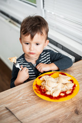 Hungry child eating dumplings in the kitchen, sitting at the table in a gray jacket