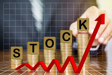 Businessman hand holding the letter stock on money coin stack arranged as a graph with red arrow on wooden table, concept of Increased investment and stock exchange