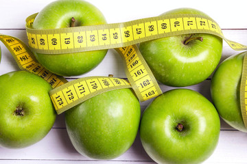 Green apples and yellow centimeter tape on white wooden background.
