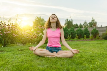 Young teenage girl practicing yoga, meditating on a summer sunset background, on green grass, lawn...