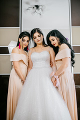Bridesmaids helping slender bride lacing her wedding white dress, buttoning on delicate lace pattern with fluffy skirt on waist. Morning bridal preparation details newlyweds. Wedding day moments.