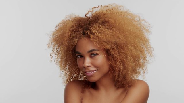mixed race woman with big curly afro blonde hair touches it and smiling to camera. Has metallic stars makeup