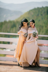 Two beautiful ladies in elegant evening dresses stand at restaurant terrace behind mountains. Beauty, fashion. Wedding style.