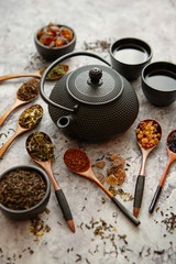 Iron vintage teapot and cups. Dried various kinds of tea on wooden spoons. Stone background with...