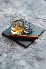 Glass of whiskey with ice cubes and cigar placed on top of stone serving plate. Stone marble...