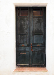 old wooden black double doors with chipped flaking faded peeling paint and rusty handles lock and letterbox in a white painted wall and tile doorstep