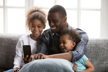 Fototapeta premium Happy African family spending time have a fun together using mobile phone at home. Black father with little daughter and son make selfie photo picture watching video sitting on couch at sitting room