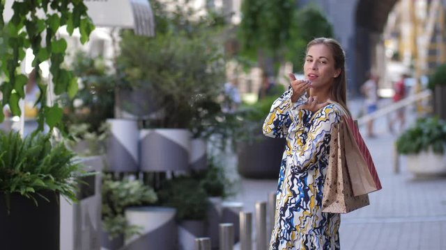 Beautiful girl looks adorable posing with shopping at the street. Woman is looking at the camera and send an air kiss. 4k slow motion