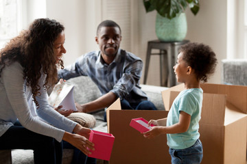 Black happy family sitting together in living room with cardboard boxes. African adorable son and laughing mother hold red gift box. Happiness moving relocate at new house, mortgage and loan concept