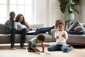 Happy black family spend free time together in living room at home. Couple sitting on sofa looks at...