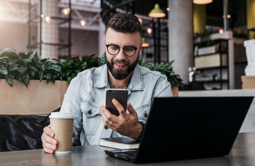 Bearded businessman sitting in cafe, talking on smart phone, working on laptop, freelancer working in coffee shop,