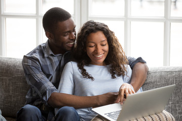 Cheerful black African married couple in love at home or hotel room. American wife husband hugging sitting on couch using computer looking on screen smiling feels happy spend time together on weekend