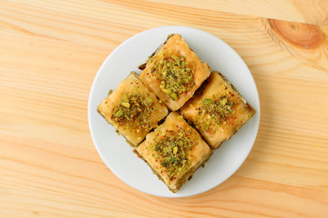 Top view of mouthwatering Baklava pastries topped with pistachio nuts on a white plate served on light brown wooden table 
