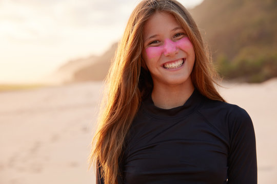 Positive young experienced surfer smiles broadly at camera, squits face with pleasure, protects face zinc dioxide, wears diving suit, ready for surfing across ocean, poses for womens magazine