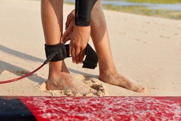 Unrecognizble barefoot woman has fixed legrope, stands on sand near surfboard, protects herself...