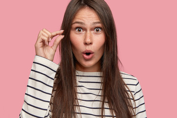 Size concept. Close up shot of amazed attractive female shapes small object with hand, demonstrates...