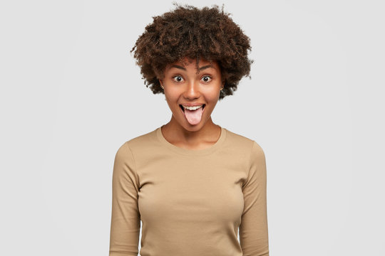 Studio shot of funny black woman shows tongue, has playful expression, Afro hairstyle, makes grimace, dressed in casual beige sweater, isolated over white background. Comic dark skinned girl