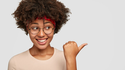 Emotive beautiful ethnic woman with pleasant smile, wears round transparent glasses, points with thumb at blank copy space for your promotional content. Dark skinned girl shows place to visit