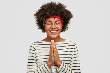 Indoor shot of happy delighted smiling woman has much dreams, poses in praying gesture as makes...