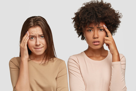 Sad dejected stressful young women have headache, keep hands on foreheads, have displeased expressions, dressed casually, isolated over white background, feel down, press lips. Horizontal shot