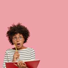 Photo of surprised dark skinned schoolgirl has shocked astonished gaze upwards, keeps mouth widely...