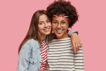 Studio shot of pretty two young happy mixed race women stand closely to each other, embrace and...