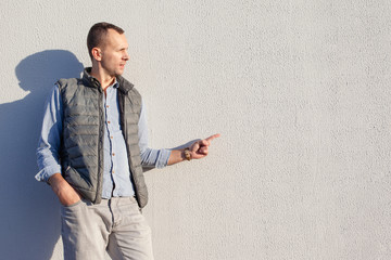 handsome young guy in gray jeans, blue denim shirt and waistcoat stands near the bright wall, copy...